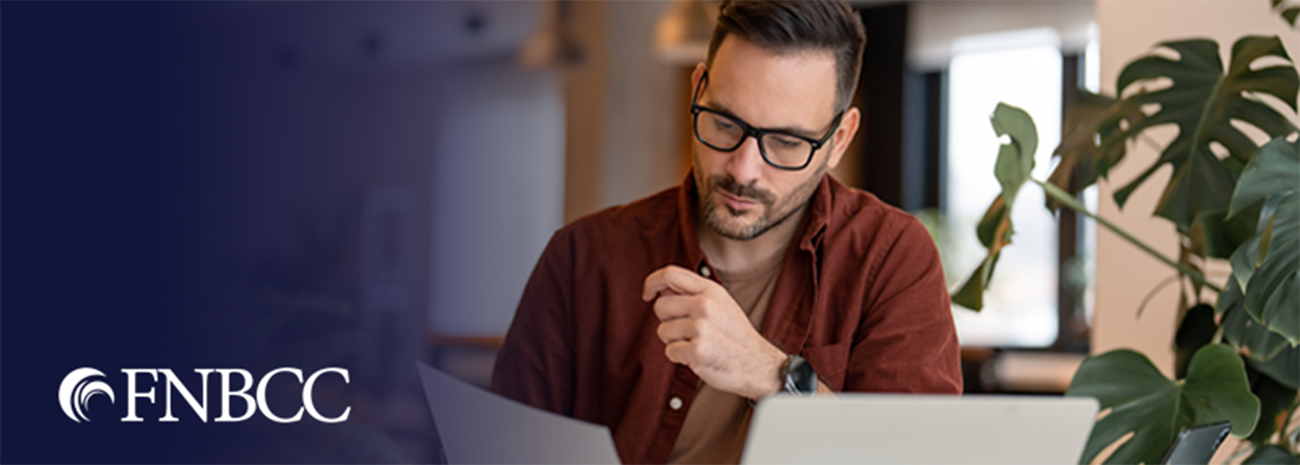 Man with glasses reviewing document in his hand while sitting in front of his laptop
